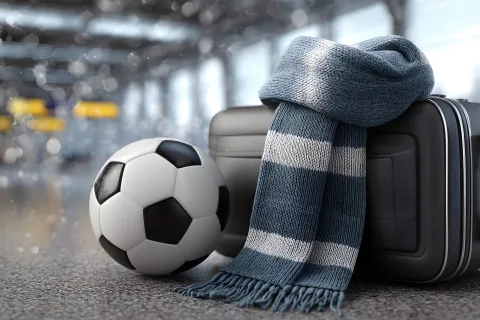 Football travel concept with soccer ball, suitcase, and winter fan scarf in an airport terminal background.