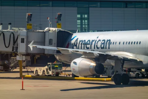 Calgary, Alberta, Canada. Nov 22, 2025. An American Airlines Airbus A320 is serviced by ground crew and jet bridge at a sunlit airport terminal gate.