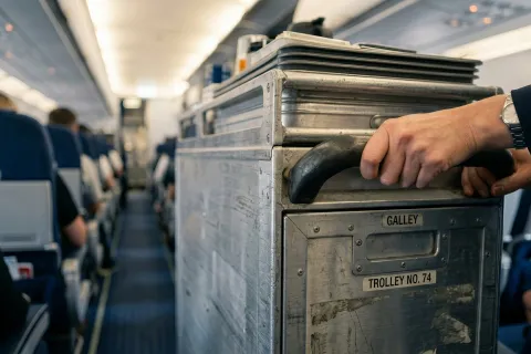 The metal catering service cart being pushed by a flight attendant inside a commercial airplane cabin.