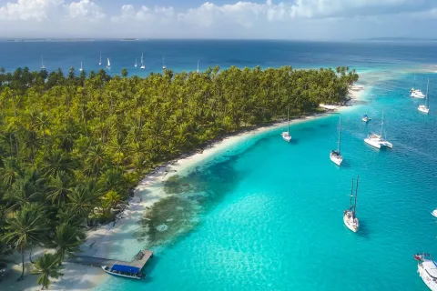 San Blas Islands, Panama - Drone Aerial View of many Sailboats & Sailing Yachts anchored in turquoise Water of Blue Lagoon next to white Sand Beach of Tropical Caribbean Island with green Palm Trees. 
