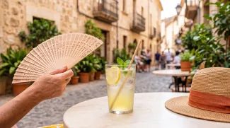 Cold lemonade with mint, straw hat, and hand fan on outdoor cafe table during European heatwave in a historic Mediterranean street.