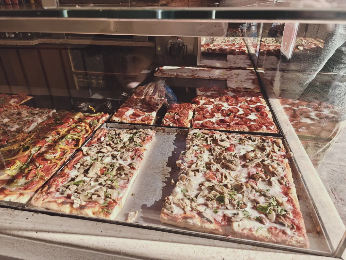 A display case full of Pizza al Taglio pick your pizza and tell them how many grams. 