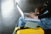 Traveler holding a passport while using a laptop to check train schedules beside a yellow suitcase